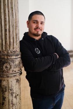 Portrait of artist Na'ye Perez wearing a black hoodie and jeans, with his arms crossed and leaning against a brown, distressed column.